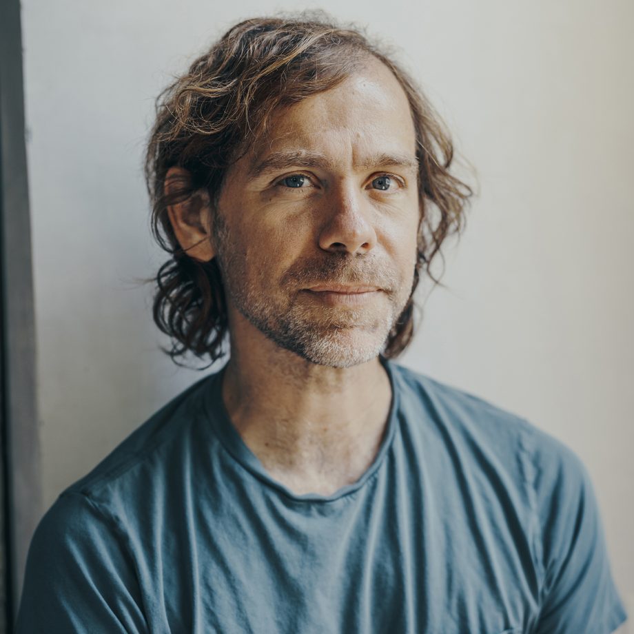 Aaron Dessner's headshot. He has shoulder length hair, is smiling slightly with his lips closed, and is wearing a blue shirt.
