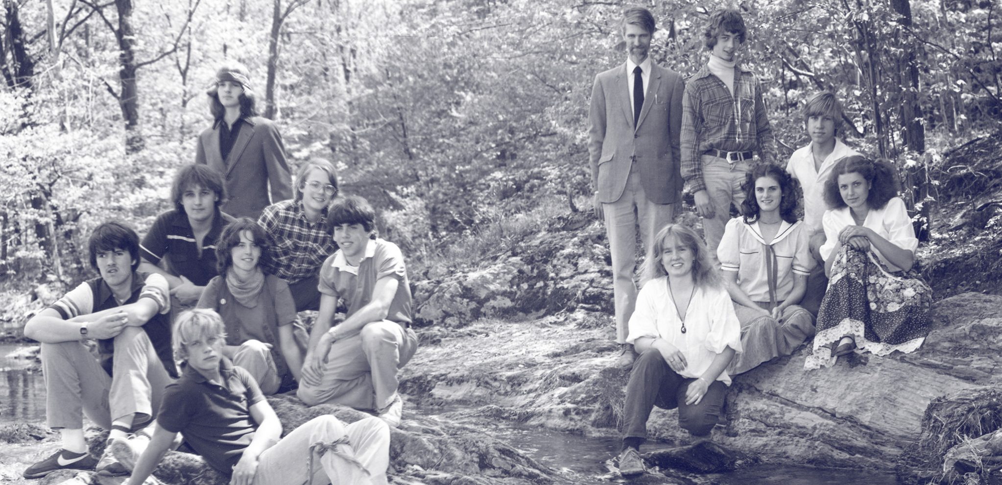 Class of 1984 and 1983 sitting in two groups outside on rocks next to the creek