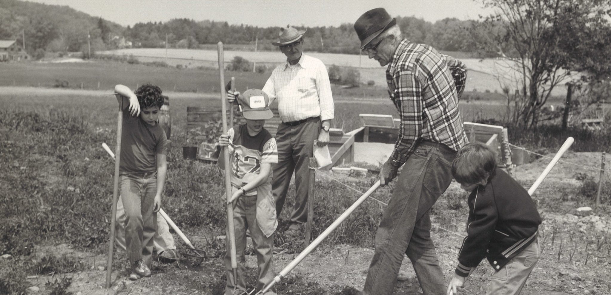 Historic photo of Fentress Gardner - one of the pioneers of Hawthorne Valley showing young students how to prepare the soil for planting; they are working with hoes and shovels with the fields behind them