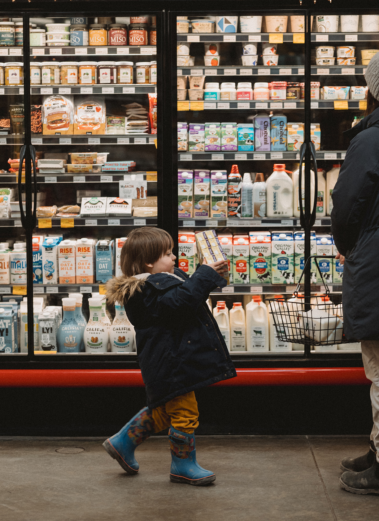 focus on a child trailing behind their mother in front of the dairy case holding a package of yogurt