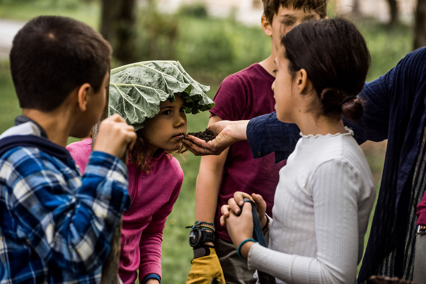 an adult holds their hand filled with dirt out to a child, wearing a giant leaf on their head, as she sniffs it and three other children are gathered around to watch